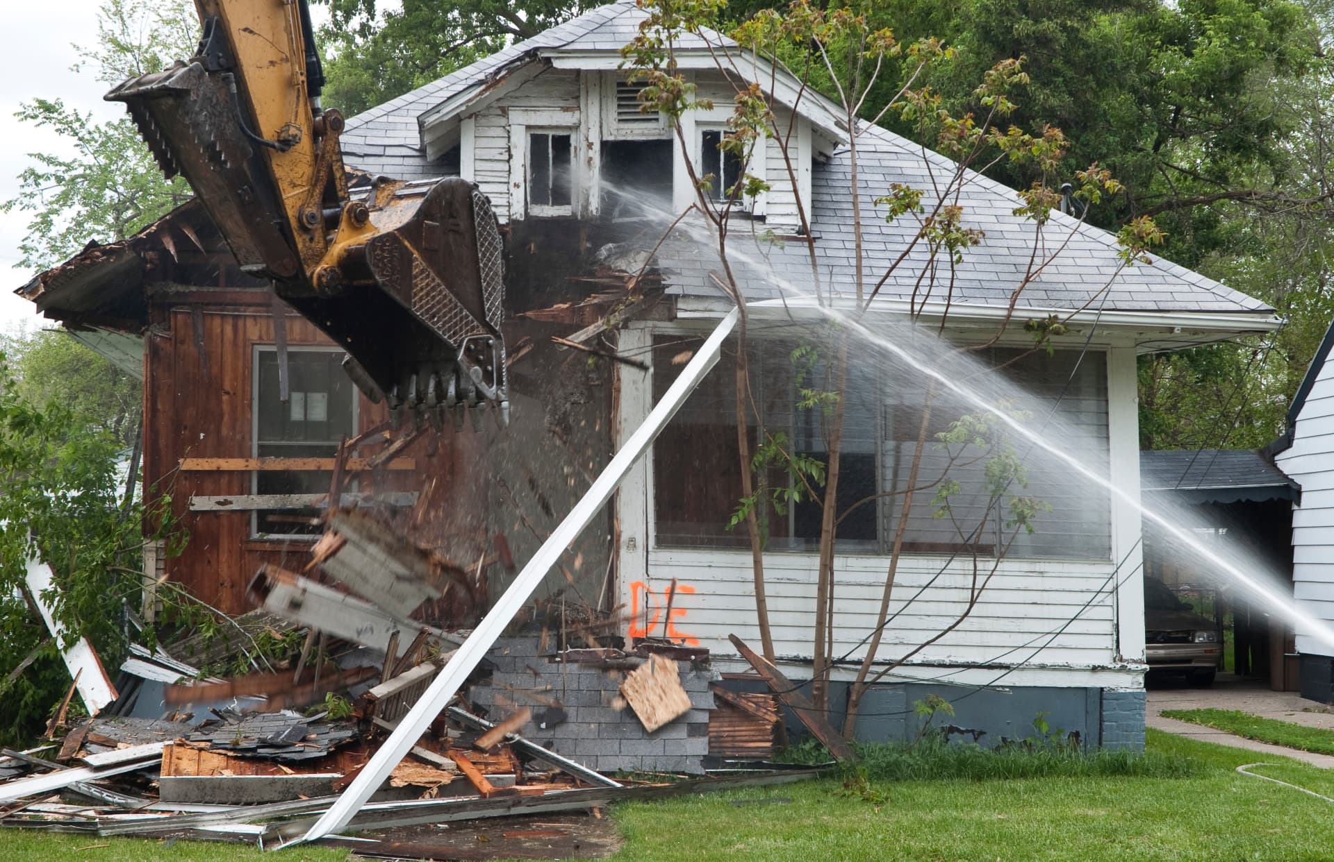Excavator taking down a house while a hose sprays the debris